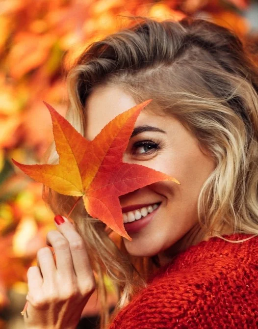 Smiling woman holding large red and orange maple leaf partially covering her face.