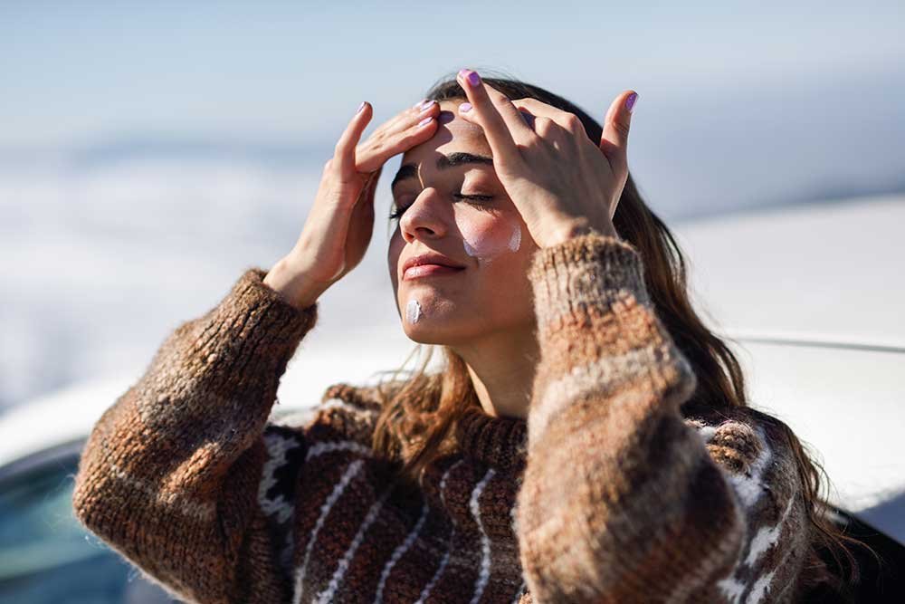 Sweater-wearing woman applying sunscreen on a bright winter day.