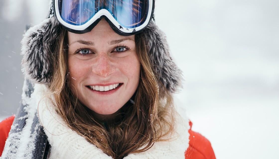 Smiling woman with skis on shoulder and fur-lined hood and googles on her head.